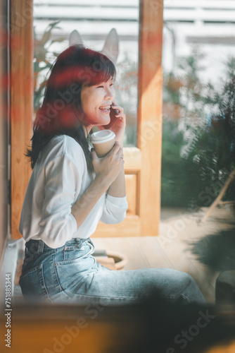 A joyful woman shares a laugh on the phone, her delight evident in her expression, as she enjoys a warm beverage by a sunlit window, creating a scene of comfort and genuine connection.