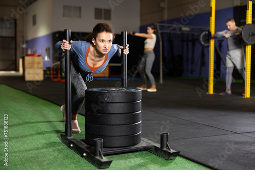 Fitness young girl pushing sled at modern gym, cross exercise with equipment during CrossFit workout photo