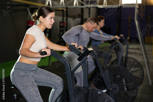 Concentrated young fit girl intensely working out on stationary air bicycle in fitness gym. Healthy active lifestyle concept..