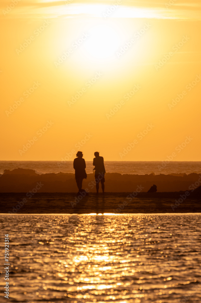 Man and woman staying and looking at sunset on ocean beach, orange sky, silhouettes of people on vacation