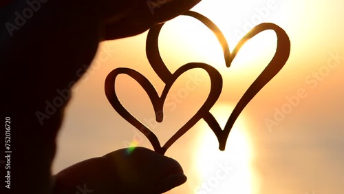A woman holding in fingers a black outline in the shape of two hearts against the background of the sun at dawn and sunset on the sea. Concept love infatuation romantic Valentine's Day mood emotions