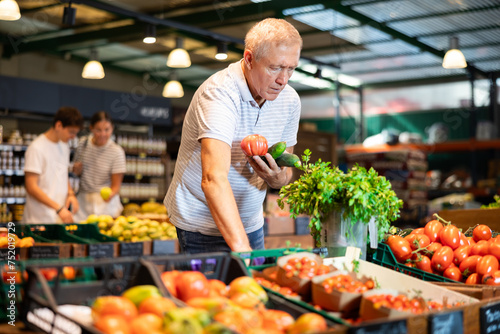 Health conscious elderly man following vegetarian lifestyle buying fresh vegetables in local grocery store, choosing ripe tomatoes and cucumbers for meal..