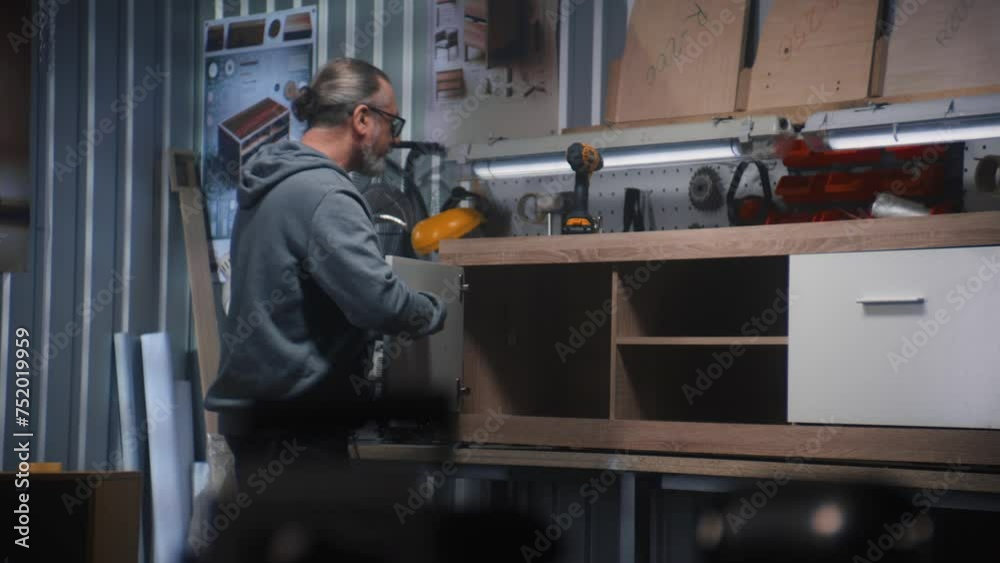 Mature carpenter in glasses makes and assembles TV stand using screwdriver. Professional male craftsman, artisan works in modern workshop for making furniture. Handicraft and small business concept. Mature carpenter in glasses makes and assembles TV stand using screwdriver. Professional male craftsman, artisan works in modern workshop for making furniture. Handicraft and small business concept.