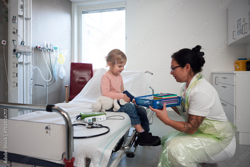 Female pediatrician giving toys to girl sitting on bed in ward at hospital Female pediatrician giving toys to girl sitting on bed in ward at hospital
