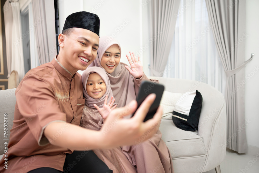 Young Indonesian family doing video call to other family and relatives with greetings gesture to the phone in happy during Eid mubarak in the living room at home.
