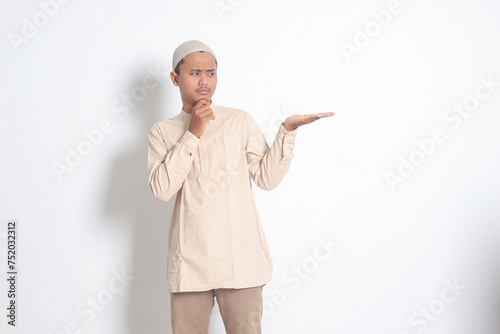 Portrait of shocked Asian muslim man in koko shirt with skullcap showing product and pointing with his hand and finger to the side. Advertising concept. Isolated image on white background