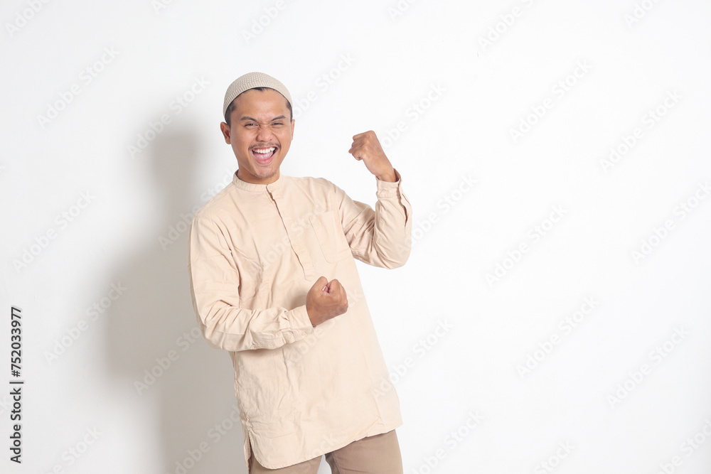 Portrait of religious Asian muslim man in koko shirt with skullcap raising his fist, celebrating success. Achievement concept. Isolated image on white background Portrait of religious Asian muslim man in koko shirt with skullcap raising his fist, celebrating success. Achievement concept. Isolated image on white background