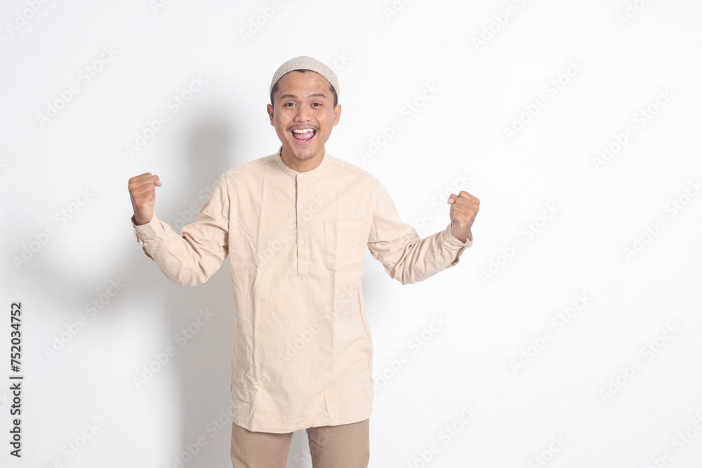 Portrait of religious Asian muslim man in koko shirt with skullcap raising his fist, celebrating success. Achievement concept. Isolated image on white background Portrait of religious Asian muslim man in koko shirt with skullcap raising his fist, celebrating success. Achievement concept. Isolated image on white background