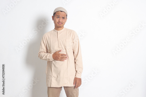 Portrait of religious Asian muslim man in koko shirt with skullcap feeling pain in his stomach, endure hunger. Stomachache and hungry for food concept. Isolated image on white background