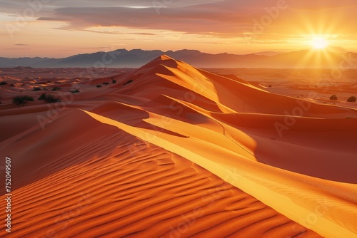 The golden hues of sunset cast over the smooth undulating sand dunes of the Sahara Desert with distant mountains silhouetted against the sunny sky