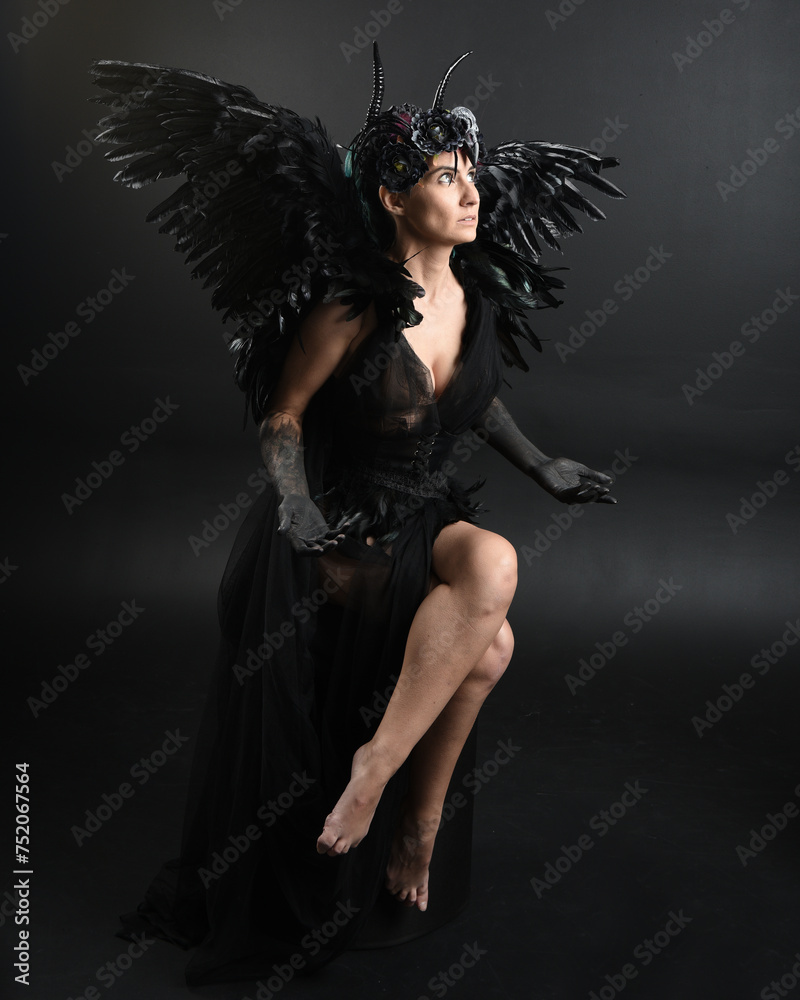 Full length portrait of model wearing gothic horned headdress with halloween black dress, fantasy angel feather wings. Crouching pose. Isolated dark studio background, cinematic shadow silhouettes 