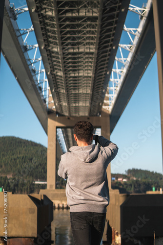 Sunny afternoon underneath Rande Bridge. Young man in a gray hoodie captures the scene with his camera, revealing the bridge's pillars over the tranquil sea, and lush green hills on the other side. photo