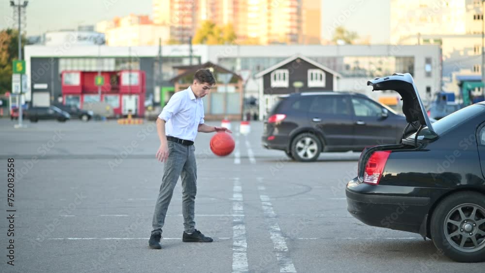 A young man in office clothes at the end of a work or school day plays with a basketball in the parking lot, throwing the ball into the trunk of a sedan A young man in office clothes at the end of a work or school day plays with a basketball in the parking lot, throwing the ball into the trunk of a sedan