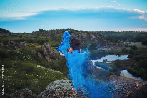 Fototapeta Naklejka Na Ścianę i Meble -  Beautiful young woman in Nature with Blue Smoke in the Air. The light and the shadow create a dramatic effect. The person is holding a blue smoke bomb.