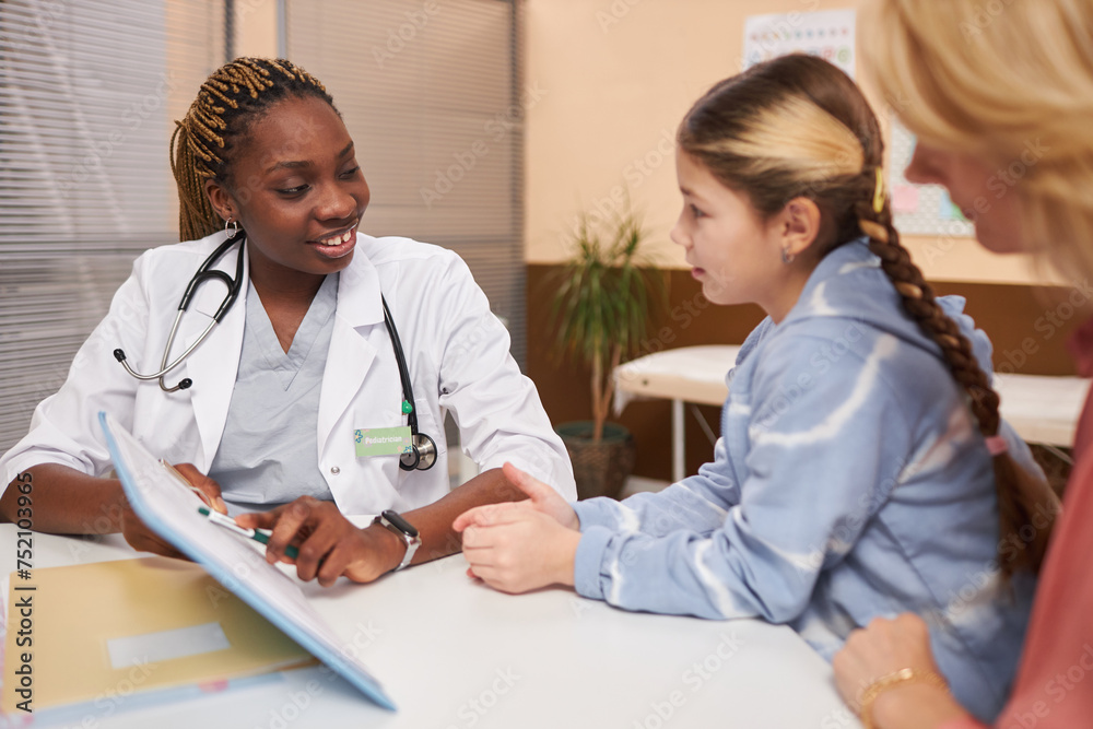 Side view portrait of smiling female pediatrician pointing at clipboard while consulting child in medical clinic and asking questions Side view portrait of smiling female pediatrician pointing at clipboard while consulting child in medical clinic and asking questions