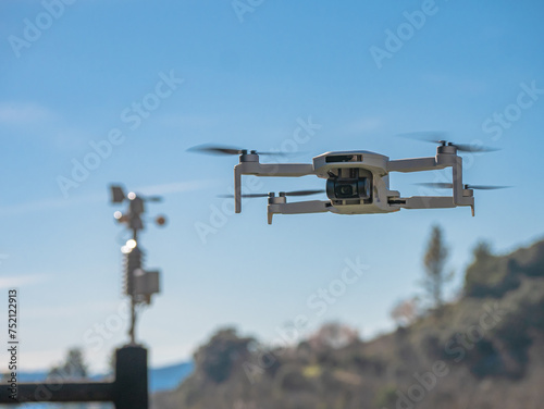 Small white drone with a built-in camera flies over a small weather station that shows the direction of the wind located on top of a wooden post and a blue sky in the background surrounded by mountain
