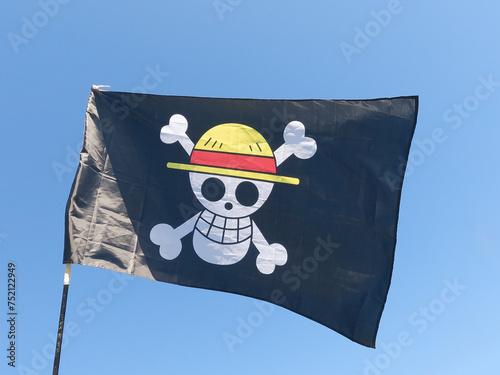 Black pirate flag which contains a white skull with a yellow straw hat and a red ribbon located on two white bones, waving in the wind on a mast and blue sky in the background