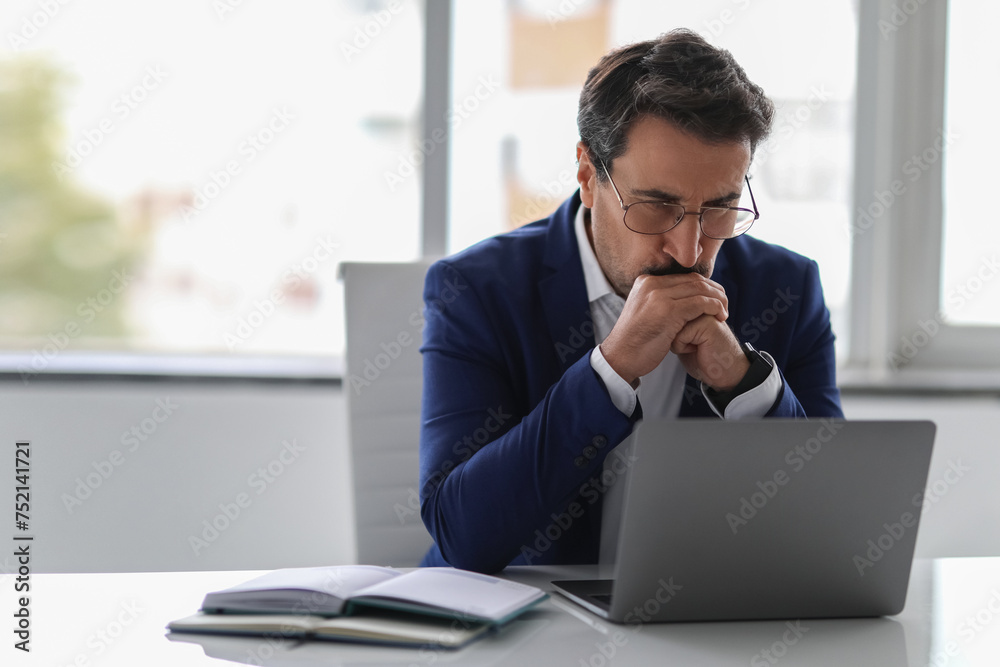 Pensive businessman in blue suit deeply focused on his laptop screen Pensive businessman in blue suit deeply focused on his laptop screen