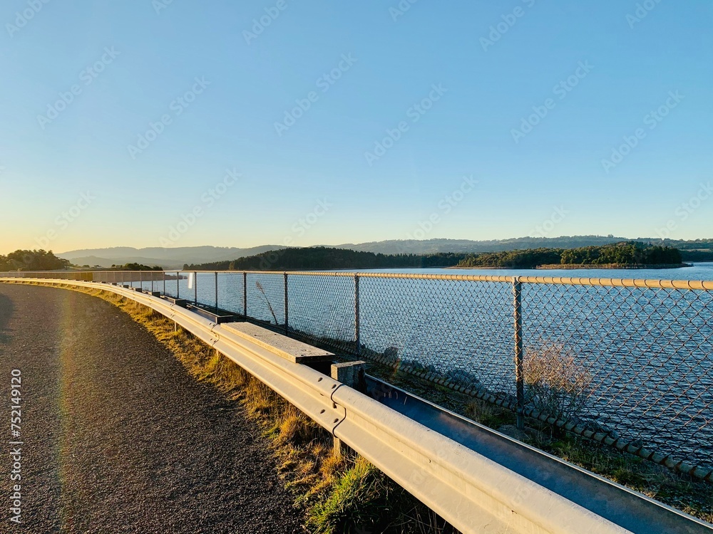 A road with a railing alongside a beautiful forest field and a serene waterscape during sunset