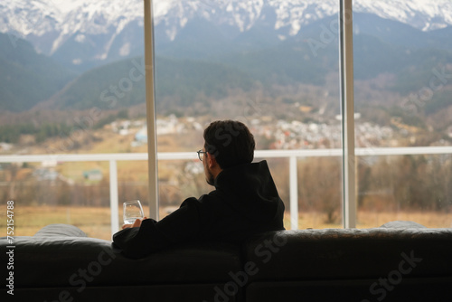 portrait of a man drinking wine while sitting on the sofa and looking at the mountains from the panoramic window of a country house