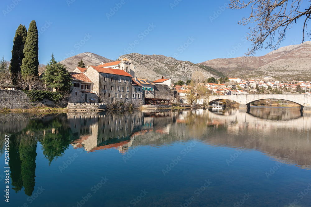 Trebinje's old town reflects in river, early springtime, bridge spans water, cypress trees, mountains behind, clear blue sky. Trebinje, Bosnia and Herzegovina