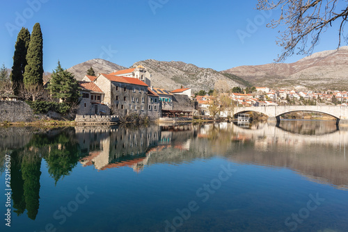 Trebinje's old town reflects in river, early springtime, bridge spans water, cypress trees, mountains behind, clear blue sky. Trebinje, Bosnia and Herzegovina photo