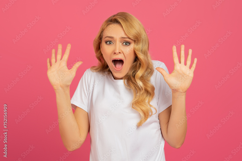 blonde woman raises hands in shocked gesture against pink backdrop blonde woman raises hands in shocked gesture against pink backdrop