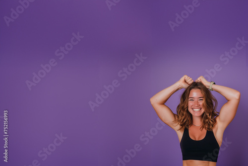 femme eurasienne très souriante, en tenue de sport qui montre ses muscles très volumineux, dans une posture de bodybuildeuse, sur un fond violet avec espace négatif pour texte copyspace.