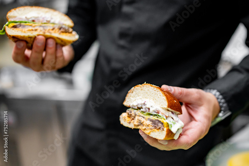 A person in black attire holds two halves of a freshly made burger, revealing grilled chicken, lettuce, and a creamy sauce. The soft bread completes this delicious meal.