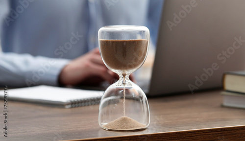 Hourglass with flowing sand on desk. Man using laptop indoors, selective focus