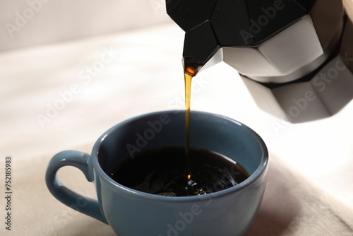 Pouring aromatic coffee from moka pot into cup at white table, closeup