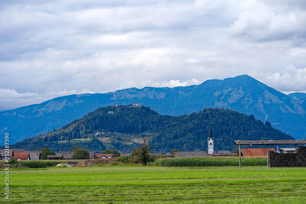 Scenic view of rural landscape at Slovenian village of Zabnica with church and mountain panorama in the background on a blue cloudy summer day. Photo taken August 10th, 2023, Žabnica, Slovenia.