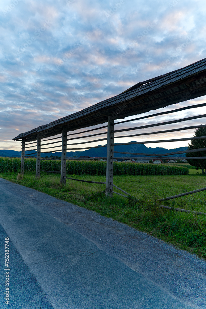 Hayrack at farmland between villages of Zabnica and Sutna with corn field and houses in the background on a blue cloudy summer evening. Photo taken August 10th, 2023, Zabnica, Kranj, Slovenia. Hayrack at farmland between villages of Zabnica and Sutna with corn field and houses in the background on a blue cloudy summer evening. Photo taken August 10th, 2023, Zabnica, Kranj, Slovenia.
