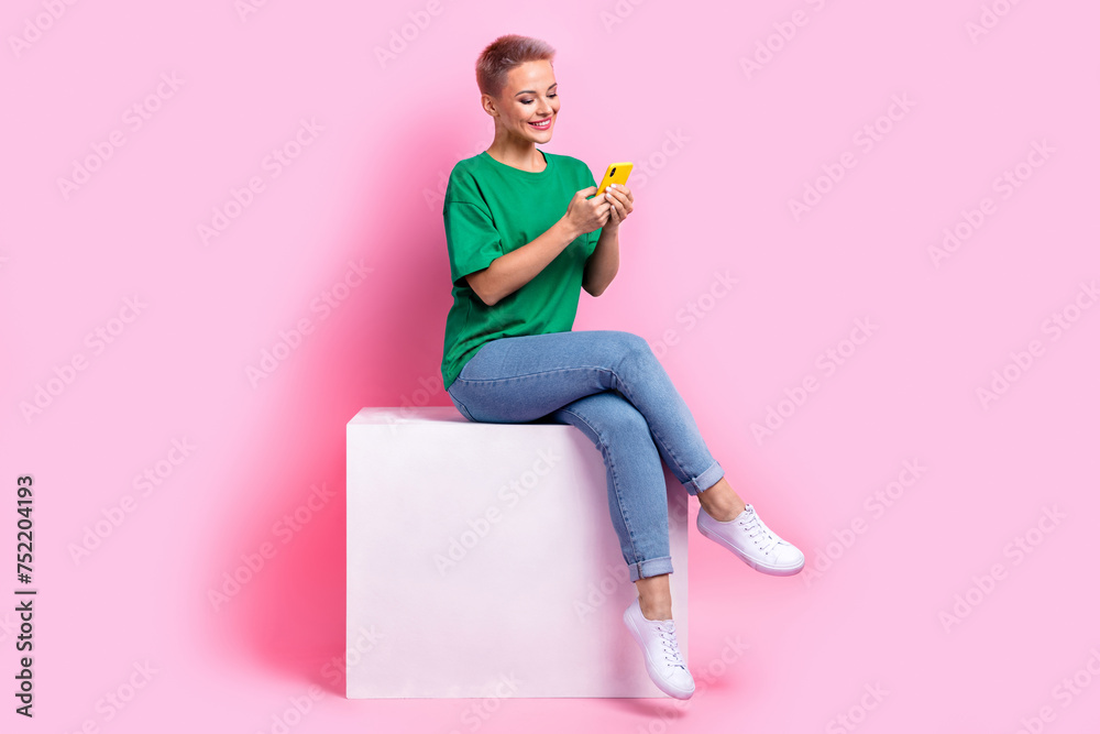 Full size photo of pleasant girl dressed green t-shirt jeans sit on white cube look at smartphone isolated on pink color background Full size photo of pleasant girl dressed green t-shirt jeans sit on white cube look at smartphone isolated on pink color background