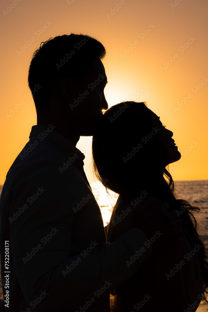 Silhouetted bride and groom against the sun with a water background.