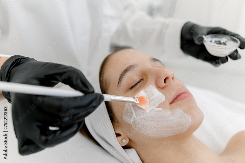 Naklejka premium The cosmetologist applies a facial white mask to the acne woman's face. Teen skin. Acne skin. A teenage girl at an appointment with a cosmetologist.