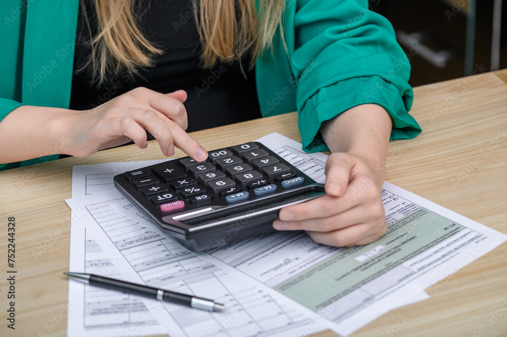 Kobieta pracująca w biurze księgowym liczy na kalkulatorze i wypełnia deklaracje podatkowe Kobieta pracująca w biurze księgowym liczy na kalkulatorze i wypełnia deklaracje podatkowe