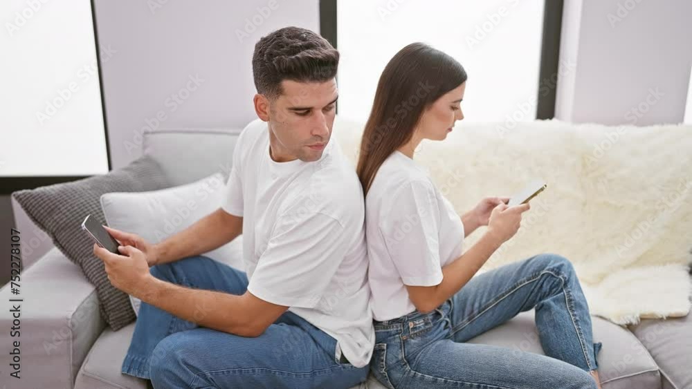 Man and woman in casual clothing using smartphones back-to-back in a modern living room, displaying disconnection despite proximity. Man and woman in casual clothing using smartphones back-to-back in a modern living room, displaying disconnection despite proximity.