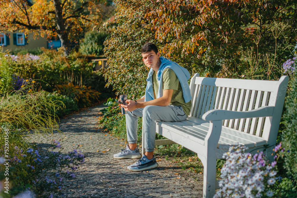 Fototapeta premium A Teenager Sits On A Bench In The Autumn Park Drinks Coffee From A Thermo Mug And Looks Into A Phone. Portrait Of Handsome Cheerful Guy Sitting On Bench Fresh Air Using Device Browsing Media Smm