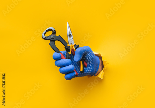 A man's hand in a blue fabric knitted glove holds wire cutters and pliers. Torn hole in yellow paper. The concept of a worker, a labor migrant, a master of his craft. Copy space. photo