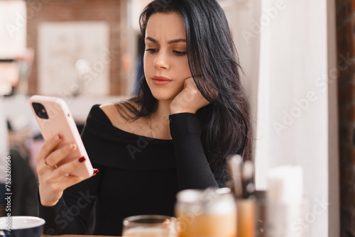 Confused puzzled brunette woman in casual clothes, sits in cafe, holds a smartphone in her hand, looks questioningly, unhappy. Girl getting surprising bad news, annoyed woman. photo