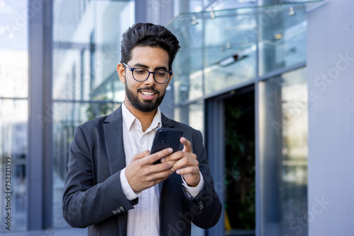 A young Indian businessman in a suit is engaged with his smartphone, smiling as he texts or reads a message. This image captures a moment of modern professional life, possibly during a break or while
