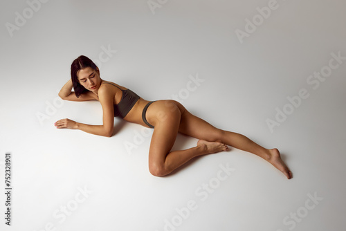 View from above of young woman posing in lingerie shows her perfect, slim body lying on floor against grey studio background. Beauty, spa procedures, dermatology treatments, cosmetology care concept.