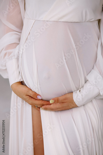 Pregnant woman in white dress holding her belly indoor. Close up photo of pregnant belly