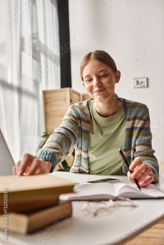 cheerful teenage girl reaching books on desk while doing homework with laptop, online class