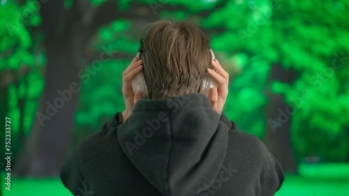 A young man enjoys music with large wireless headphones in the afternoon against the backdrop of a green park. Back view.