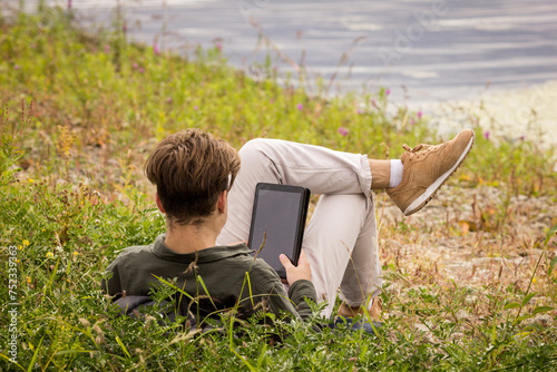 A teenager lies in the grass with a tablet in his hand