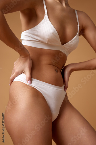 Close-up of woman in white underwear, showcasing belly with smooth and toned skin against sandy color studio background. Concept of beauty, spa procedures, dermatology treatments, cosmetology care.