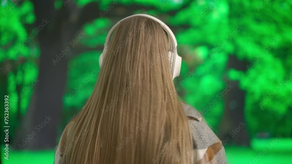 A young woman enjoys music with large wireless headphones in the afternoon against the backdrop of a green park. Back view. A young woman enjoys music with large wireless headphones in the afternoon against the backdrop of a green park. Back view.