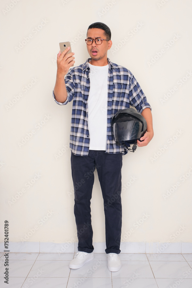 A man carrying motorcycle helmet showing shocked expression when looking to his phone A man carrying motorcycle helmet showing shocked expression when looking to his phone